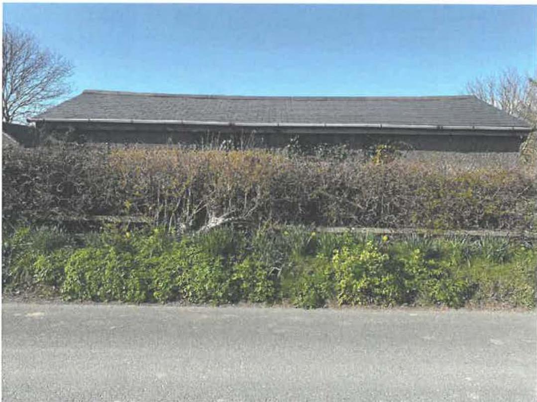 A photograph showing a long, single-story stone building with a pitched roof, likely a stable block, situated behind a dense hedge and vegetation.