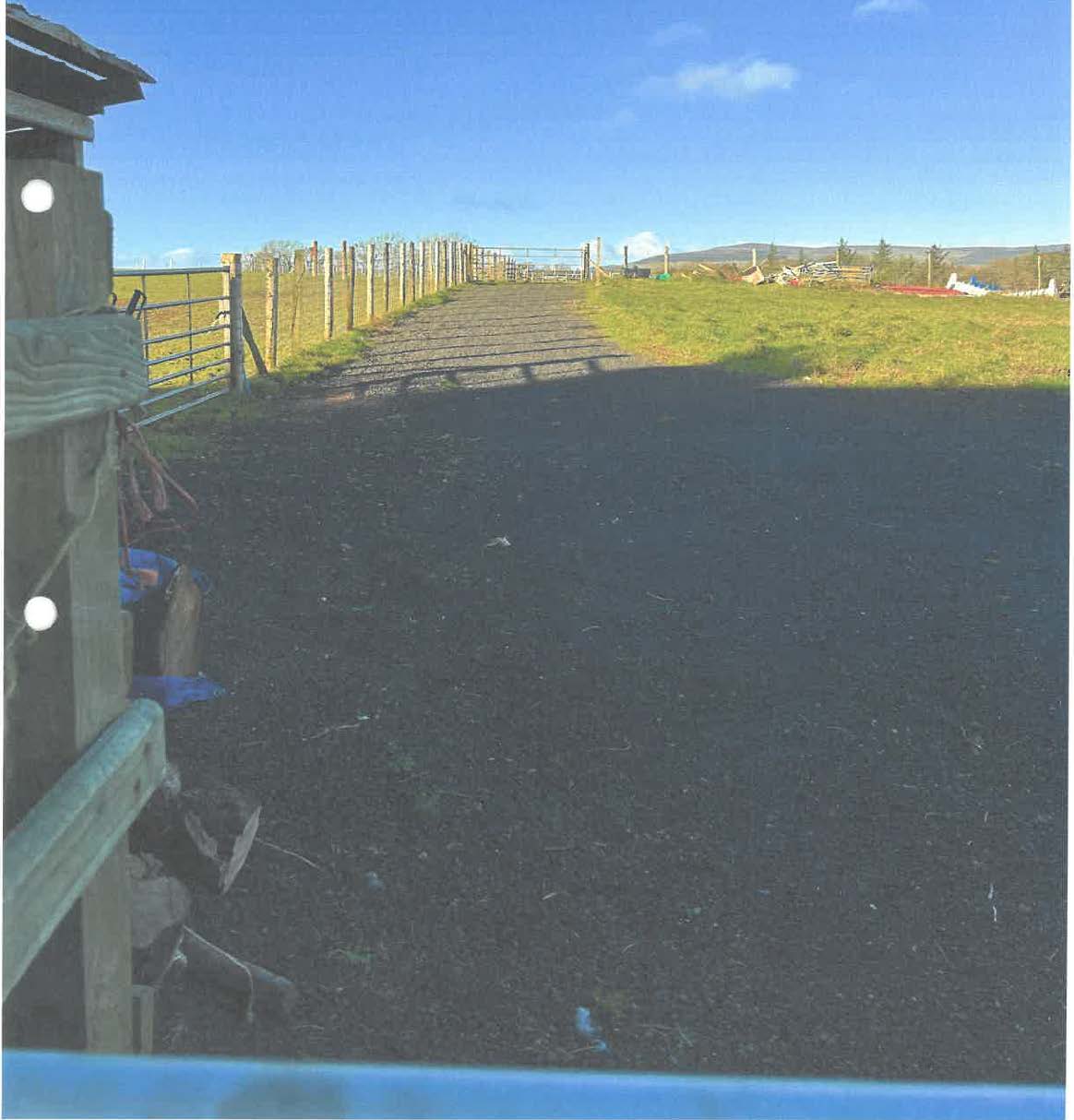 A photograph showing a rural access track or hardstanding area bordered by wooden fencing and grassy fields.