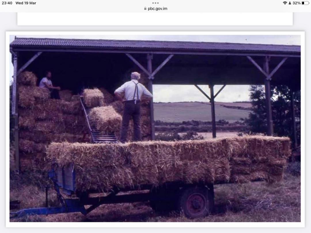 A photograph depicting a rural agricultural setting with a large open-sided barn structure and hay bales being loaded onto a trailer.