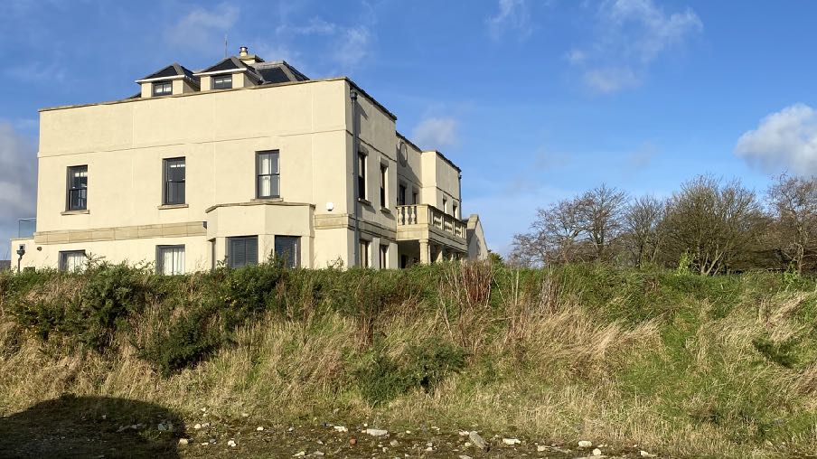 A large, cream-colored detached house situated on a grassy embankment under a blue sky.