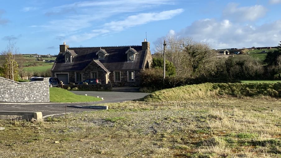 A photograph of a rural site showing an existing stone house, a newly paved driveway entrance, and a stone wall in the foreground against a backdrop of green hills.