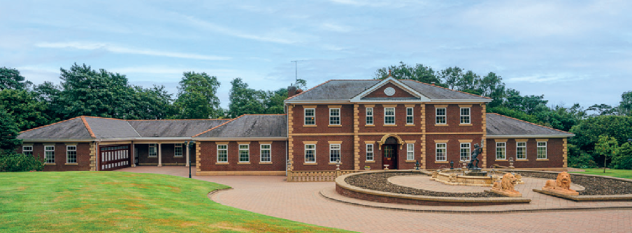 A panoramic photograph shows a large, multi-winged brick residence with a formal paved forecourt featuring a circular garden element and statues. The property is set in a rural environment with extensive lawns and mat...