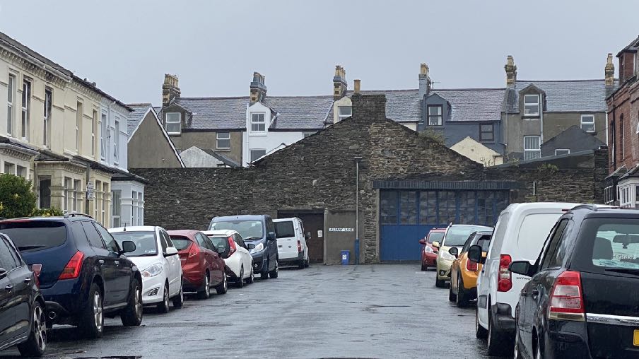 A street-level photograph showing a row of parked cars in front of a stone building with a blue garage door and a smaller pedestrian door, with terraced houses in the background.