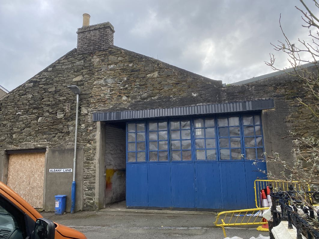 A street-level photograph of a stone building featuring a large blue garage door and a smaller boarded opening, situated on Albany Lane with construction barriers nearby.