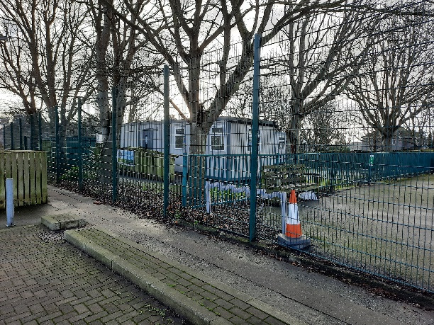 A photograph showing existing white mobile classroom units situated behind a green metal fence, viewed from a paved pathway with bare trees in the background.