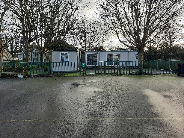 A photograph showing two white prefabricated mobile units or classrooms situated behind a green metal fence, surrounded by trees.