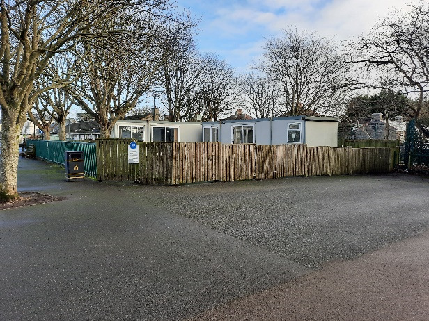 A photograph showing a row of white mobile classroom units situated behind a wooden fence, with large trees in the background and a paved area in the foreground.