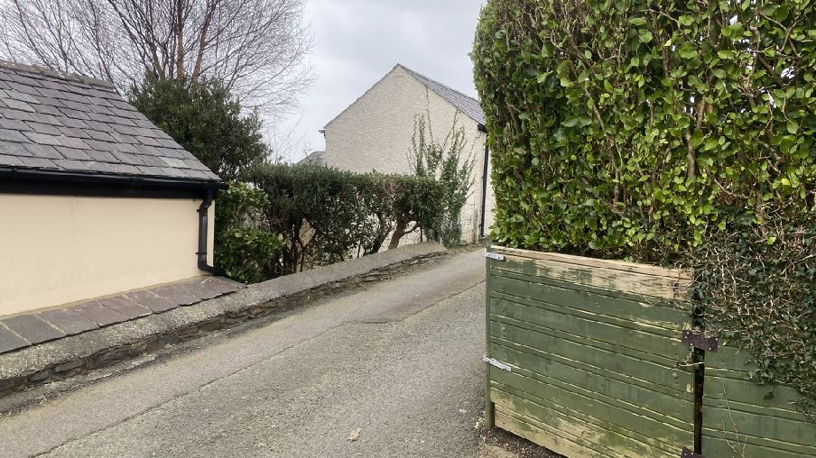A photograph showing a paved driveway or lane leading uphill, bordered by a stone wall and building on the left and a wooden gate with a hedge on the right.