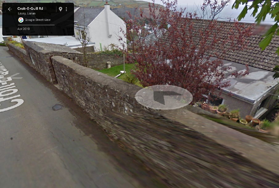 A blurry Google Street View screenshot showing a long stone boundary wall running alongside a road in a rural area with houses visible behind it.