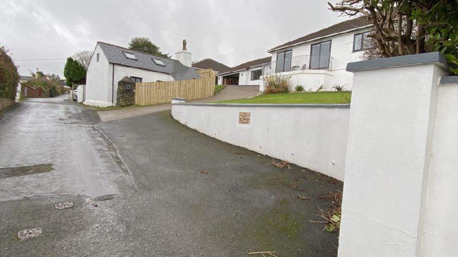 A street-level photograph showing a rural property with a white house featuring a balcony and a white retaining wall in the foreground. To the left, there is a white building with a stone wall section and wooden fenci...