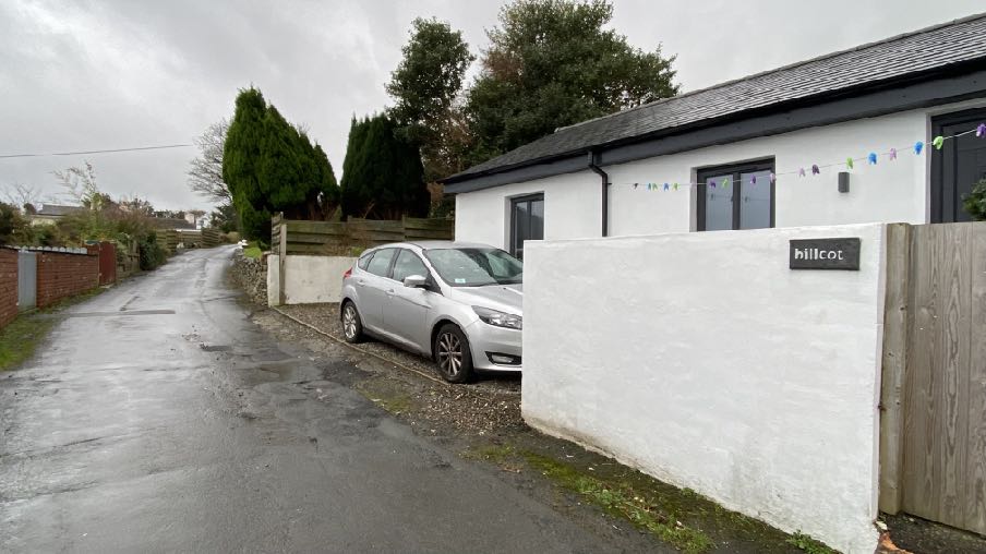 A street-level photograph showing a white bungalow with a new white rendered wall and a silver car parked on a gravel area next to a rural road.