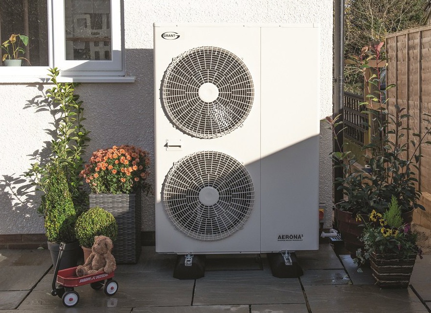 A photograph of a white Grant Aerona air source heat pump unit situated on a paved patio area next to a house wall and garden plants.