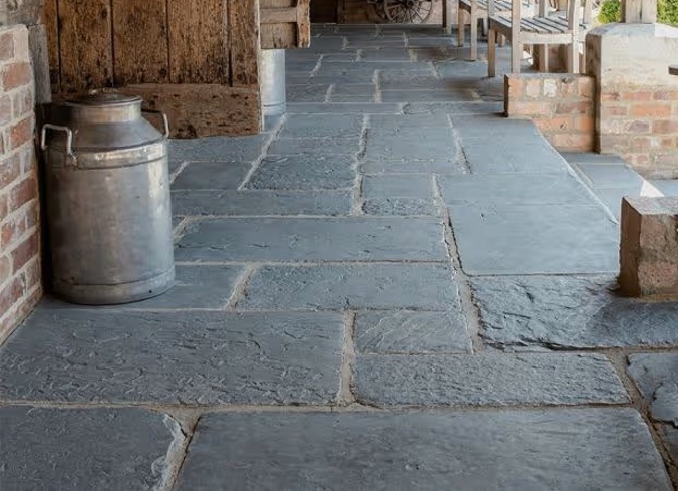 A close-up photograph showing a slate paved patio or walkway next to a brick and timber structure, featuring a large metal container.
