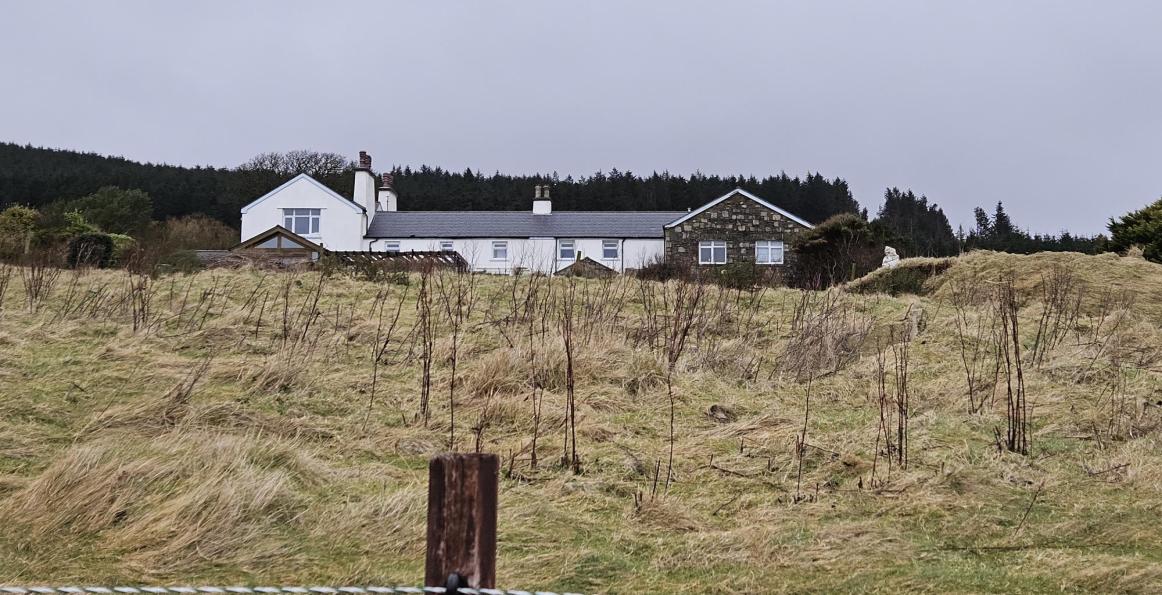 A long, single-story dwelling with mixed white and stone walls is situated on a grassy slope in a rural setting.