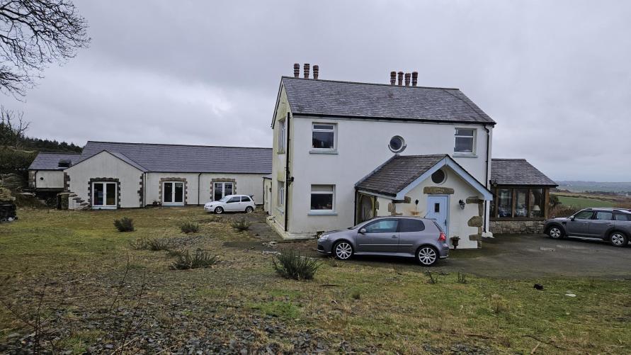 A photograph showing a white two-story detached house with a slate roof and a single-story annex or garage structure to the left, set in a rural grassy area with parked cars.