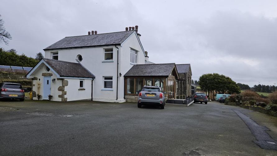 A photograph of a white two-story detached house with a slate roof, featuring a glass-fronted extension on the right and a smaller outbuilding on the left.