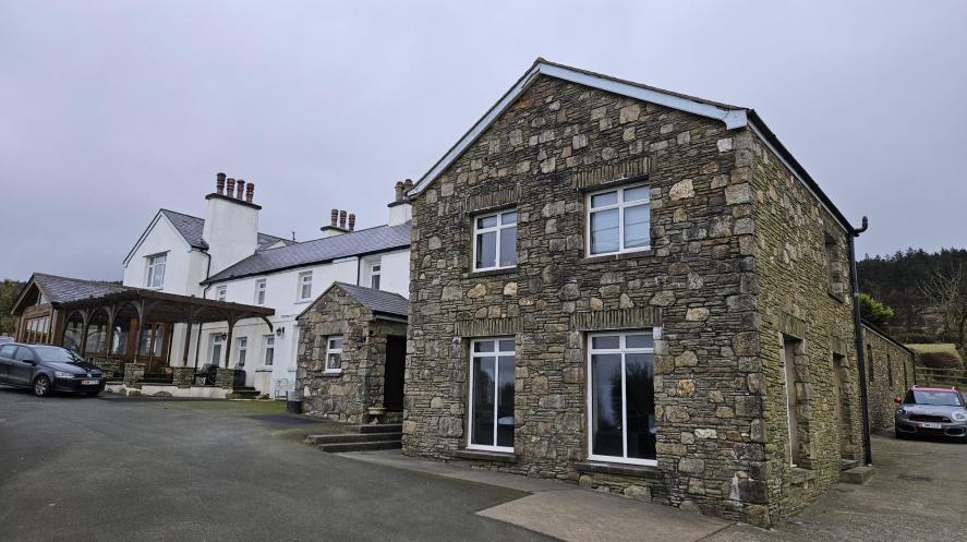 A photograph showing a detached stone building with large windows in the foreground and a white house with a porch in the background.