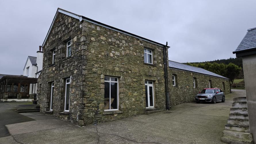 A photograph showing a two-story stone dwelling with white windows and a paved driveway, with a long single-story stone outbuilding visible in the background.