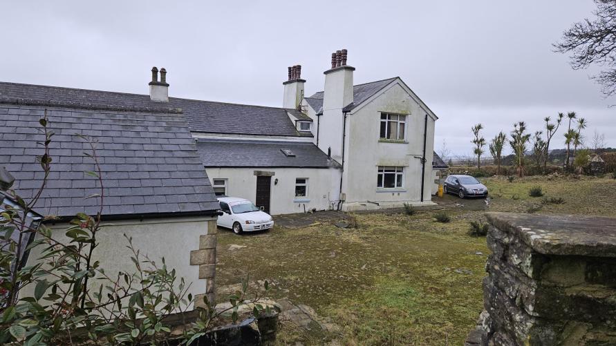 A photograph of a white, detached stone cottage with a slate roof and chimneys, set within a grassy yard with parked cars and a stone wall in the foreground.