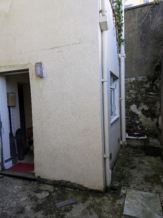 A photograph showing the exterior corner of a rendered building with a doorway and window, alongside a narrow stone-walled alleyway.