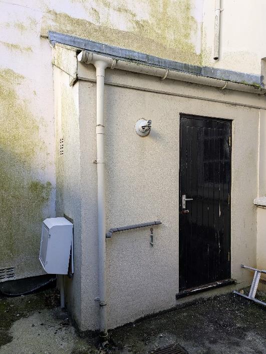 A photograph showing the exterior corner of a building with a black door, a security light, and a white downpipe running down the wall.