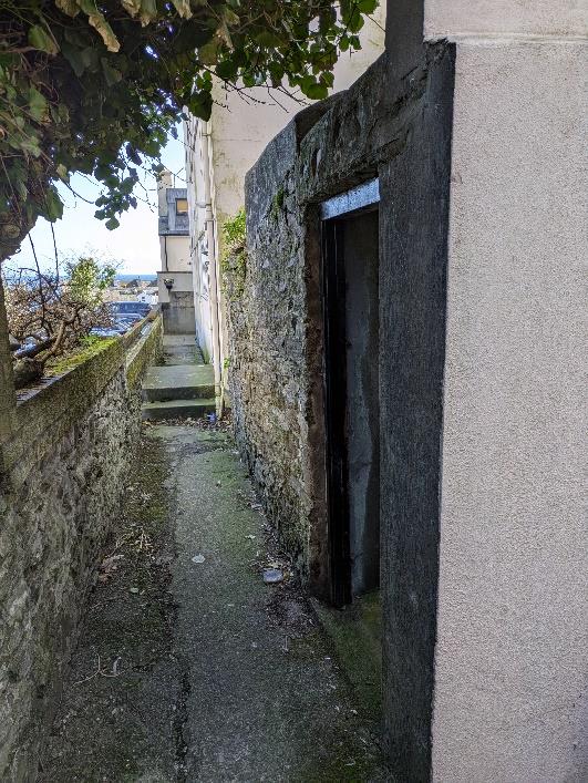 A photograph showing a narrow stone-walled alleyway or passage between buildings, with steps leading up in the background and a coastal view in the distance.