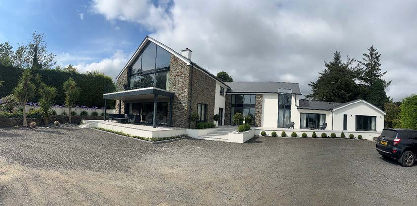A wide-angle photograph of a modern detached house featuring stone and white cladding, a large gravel driveway, and a single-story white extension on the right side.