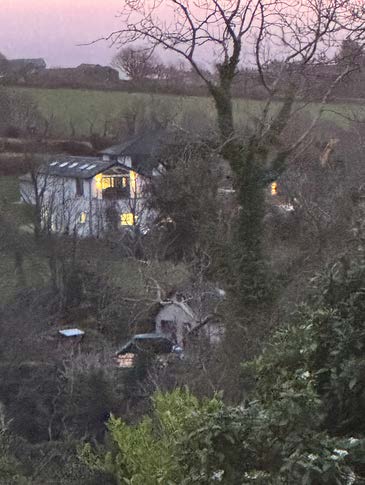 A dusk photograph showing a white house with illuminated windows in the background and a smaller outbuilding structure in the foreground surrounded by trees.
