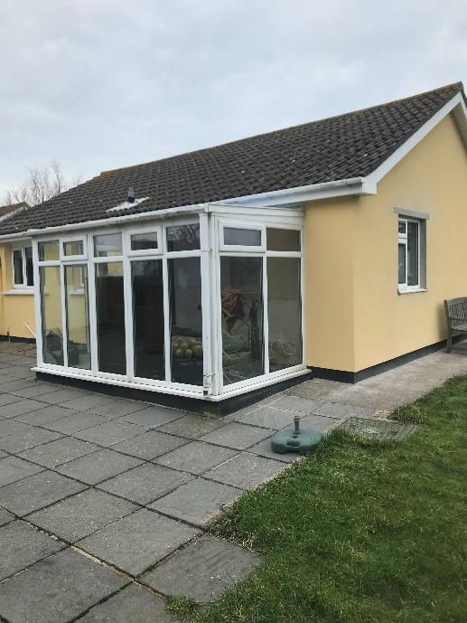 A photograph showing a single-story yellow house with a large white-framed conservatory extension attached to the side, overlooking a paved patio area.