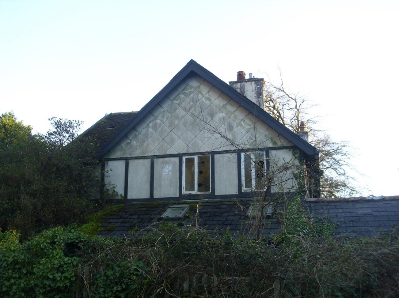 A photograph showing the upper gable end and roof of a residential building with diamond-patterned cladding and timber framing, surrounded by overgrown vegetation.