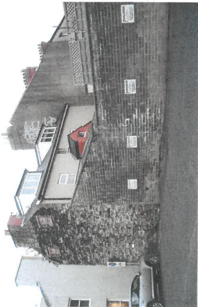 A rotated photograph showing a street scene with a brick wall featuring planning notices, a white building with a red dormer window, and an adjacent stone building.