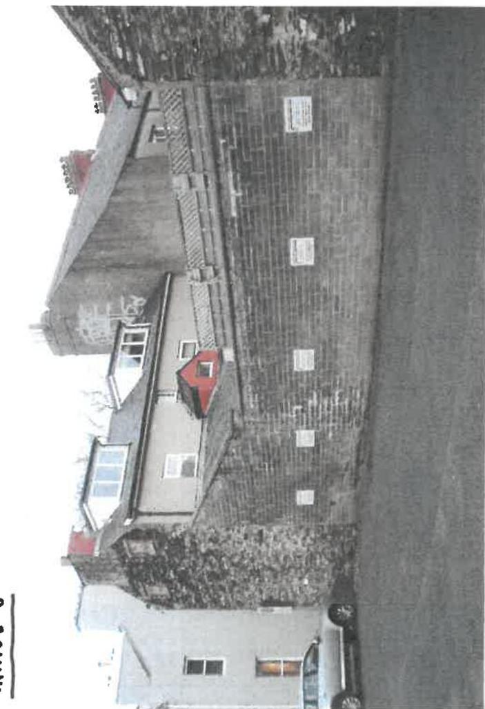 This is a rotated photograph showing the existing facade of a terraced building, likely a shop with residential accommodation above. It features dormer windows, a brick wall, and a parked car at street level.