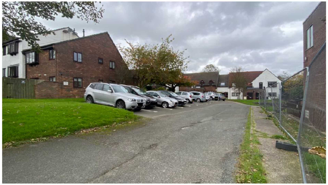 A street-level photograph showing a large brick building with a parking lot in front, likely the former nursing home mentioned in the application. The scene includes parked cars, a paved road, and surrounding resident...