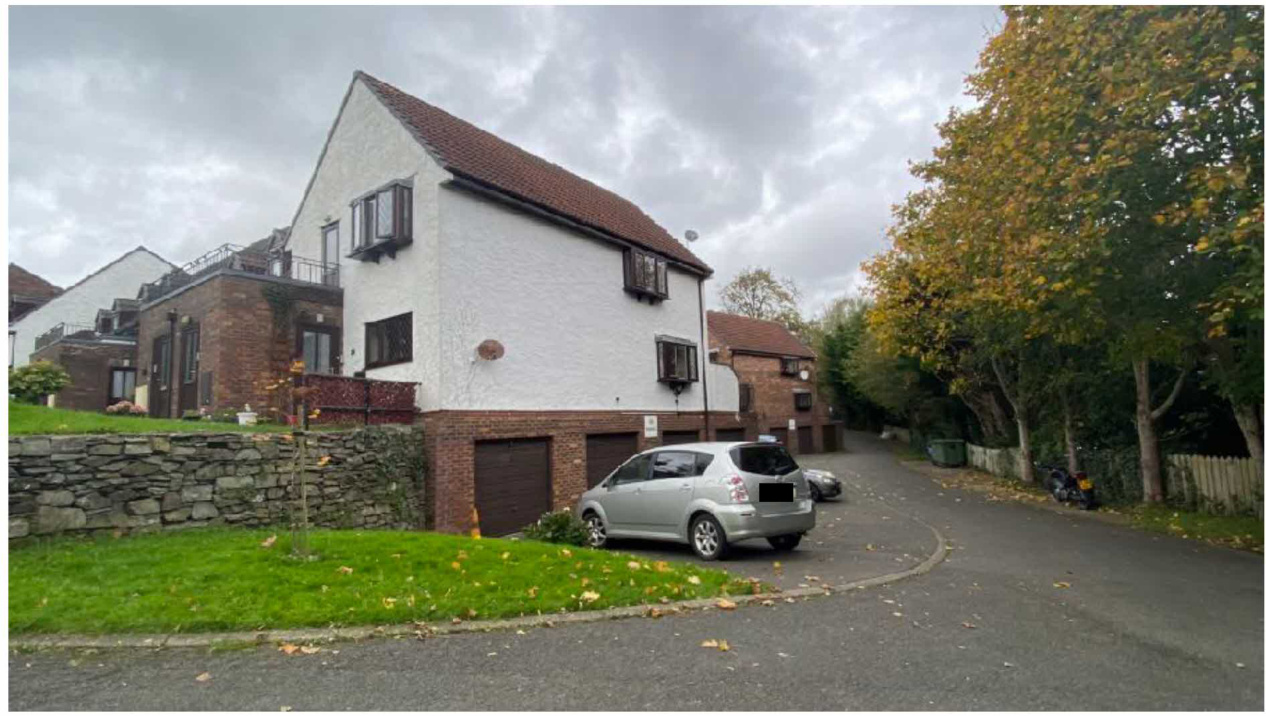 A street-level photograph showing a white two-story residential building with a brown tiled roof and garage doors, situated next to a brick building and a stone wall.