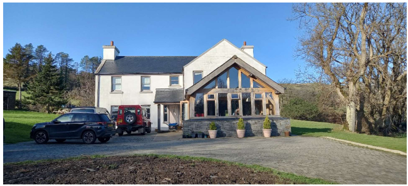 A photograph of a white detached house featuring a large timber-framed glazed extension and a gravel driveway with parked cars in a rural setting.