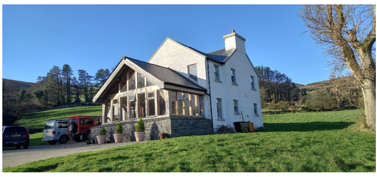 A photograph showing a white two-story house with a large modern glazed extension, situated in a rural setting with parked vehicles.