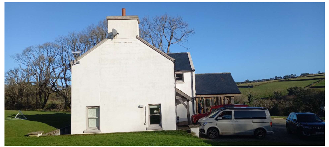 A photograph showing a white rendered building, likely a converted barn, with a slate roof and chimney stack. An attached wooden structure is visible to the right, with vehicles parked in the foreground against a rura...