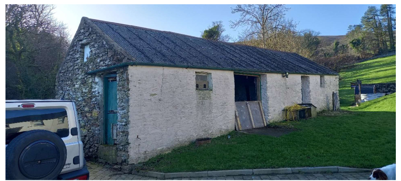 A photograph showing an existing stone and rendered barn with a slate roof, situated in a rural setting with a white vehicle parked nearby.