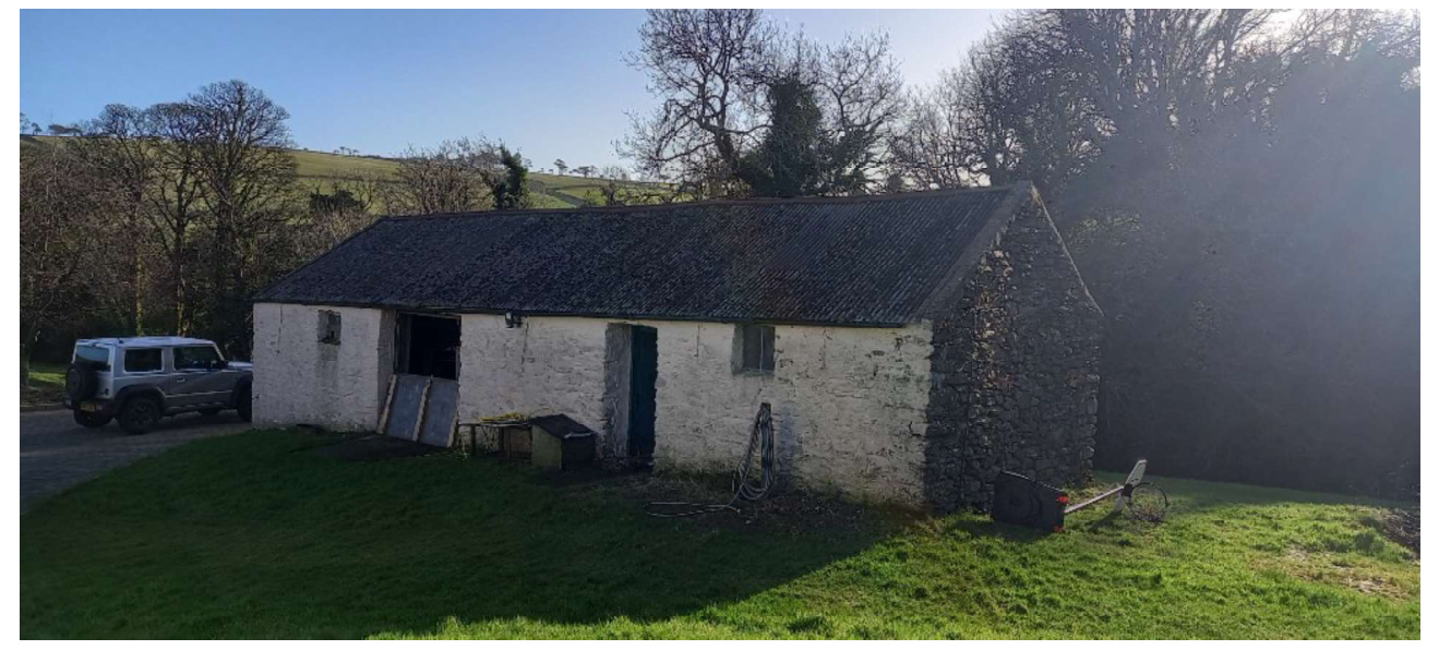 A photograph of an existing white-washed stone barn with a slate roof, situated in a rural grassy area with a car parked nearby.