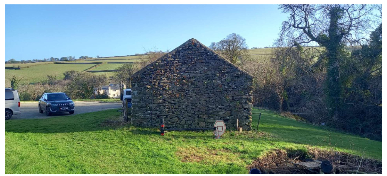 A photograph showing an existing stone barn structure in a rural landscape with vehicles parked nearby.