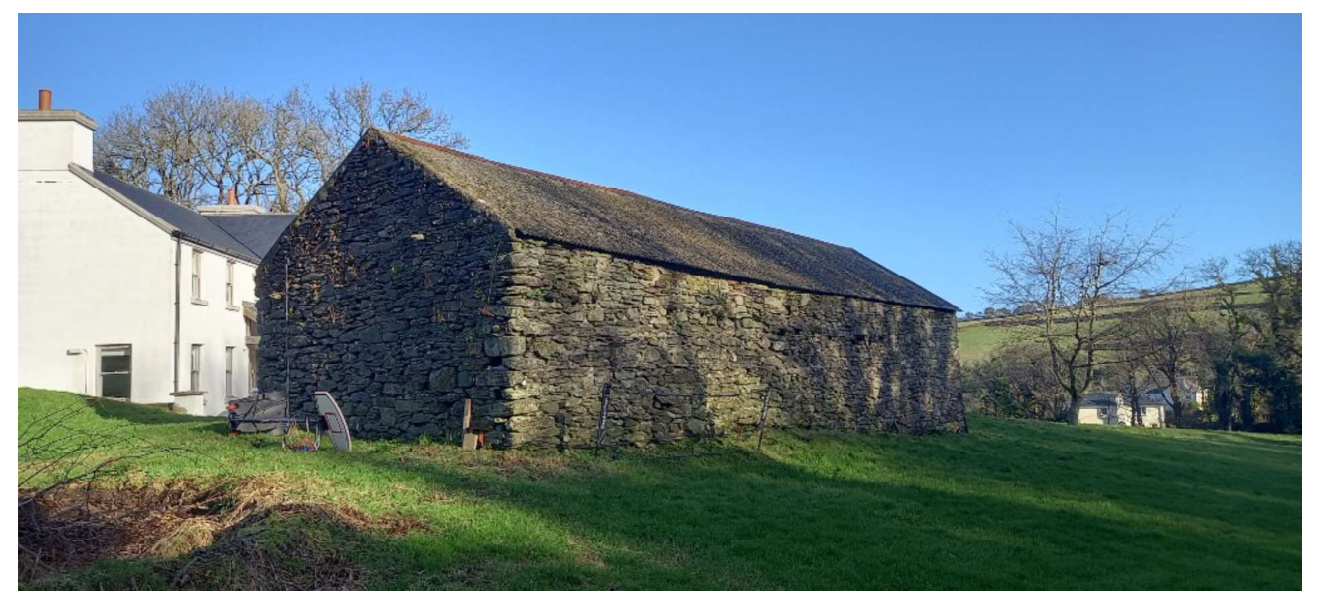 A photograph showing a large, traditional stone barn adjacent to a white residential building in a rural setting.