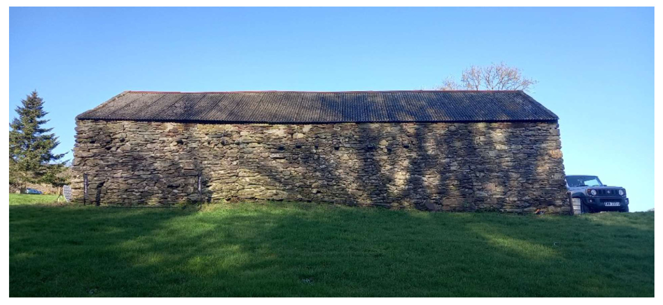 A photograph showing a long, single-story stone barn with a slate roof situated in a grassy field, with a car parked to the right.