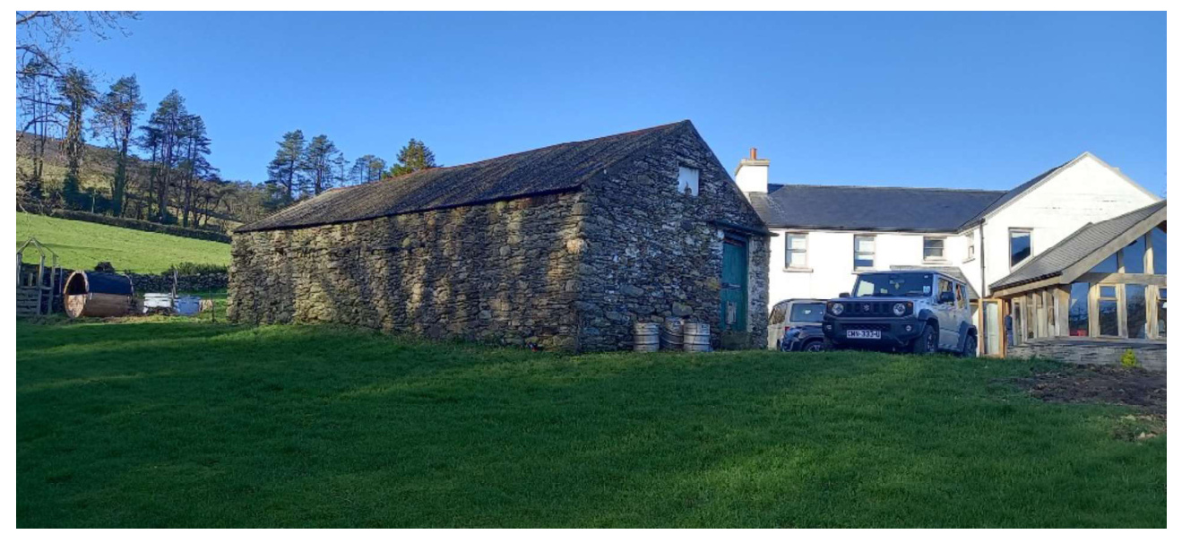 A photograph showing a large traditional stone barn situated next to a white residential house with a modern extension in a rural setting.
