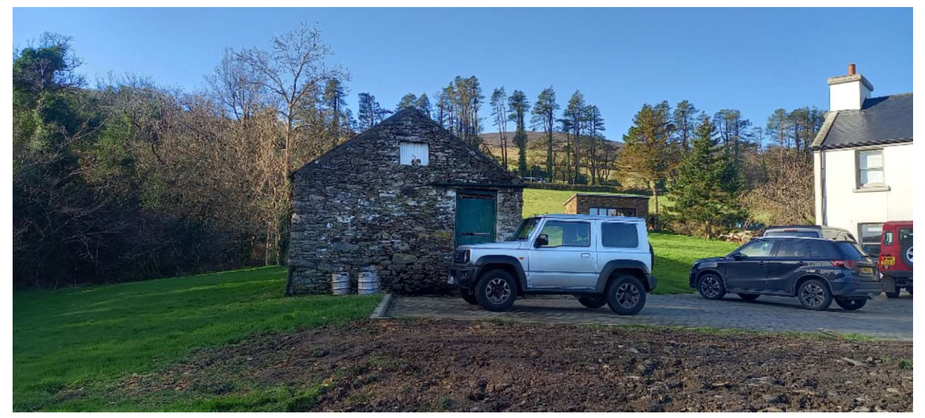 A photograph showing an existing stone outbuilding or barn situated next to a white house, with vehicles parked on a driveway in a rural setting.