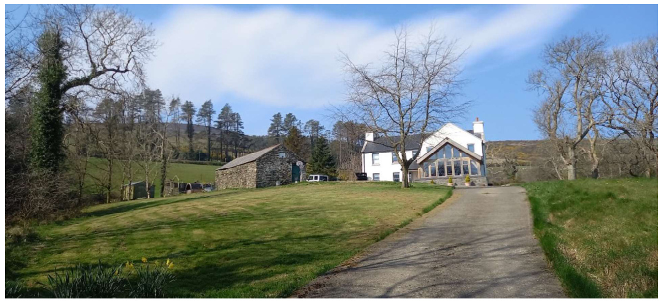 A photograph showing a rural property featuring a white house with a large glass extension and an adjacent stone barn structure, viewed from a grassy slope with a driveway.