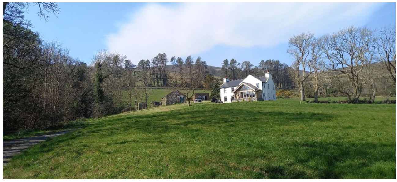 A wide-angle photograph showing a white house and a stone barn situated on a grassy slope in a rural setting with trees and hills in the background.