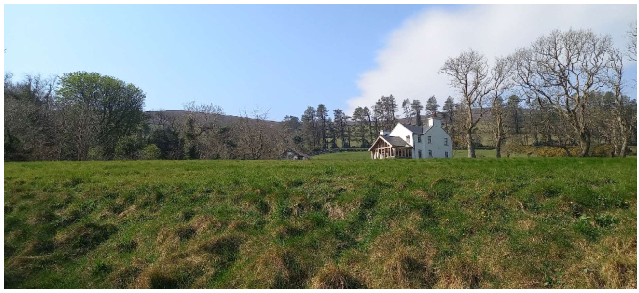 A photograph showing a white detached house with a wooden porch extension situated in a grassy rural field with trees in the background.