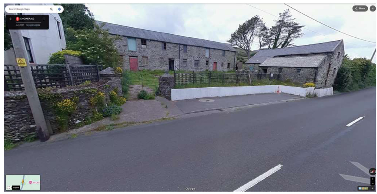 Google Street View screenshot showing stone barn structures and a white retaining wall along a rural road.