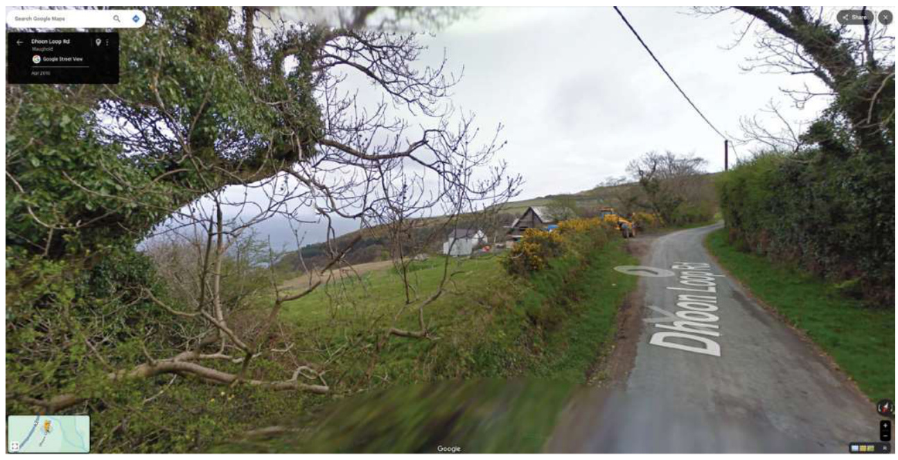 A Google Street View screenshot showing a narrow rural road named Dhoon Loop Rd in Maughold, with surrounding vegetation and distant buildings on a hillside.