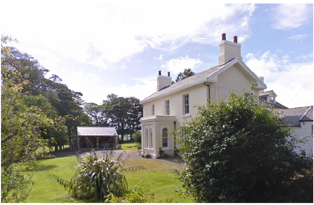 A photograph showing a large white detached house with a garden and a wooden outbuilding structure to the left.